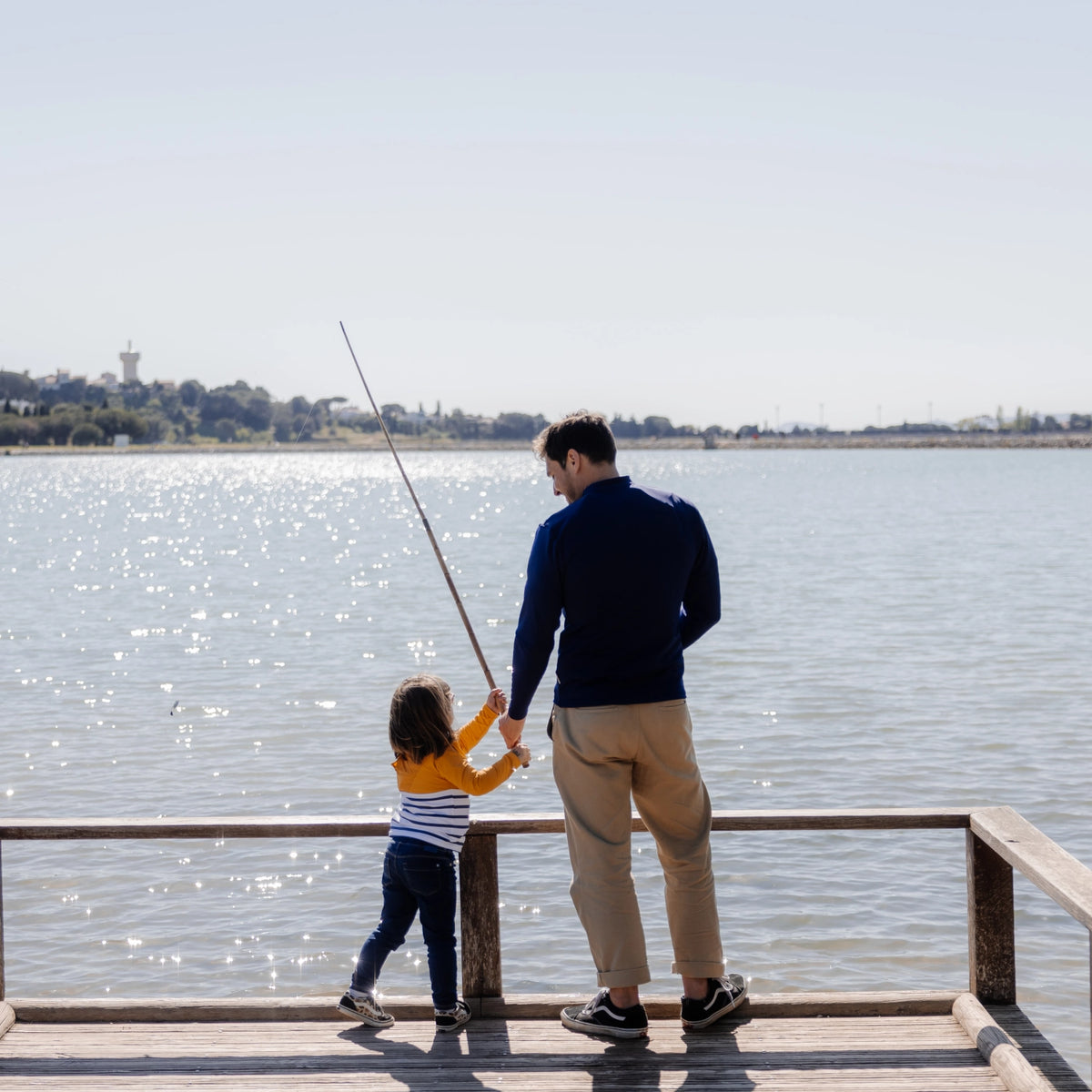sur un ponton au bord d'un lac, papa portant un T-shirt anti-noyade adulte automatique Floatee, couleur bleue et manches courtes, et petite fille portant un tee-shirt anti-noyade enfant Floatee, couleur jaune et manches longues. Les deux tiennent une canne à pêche