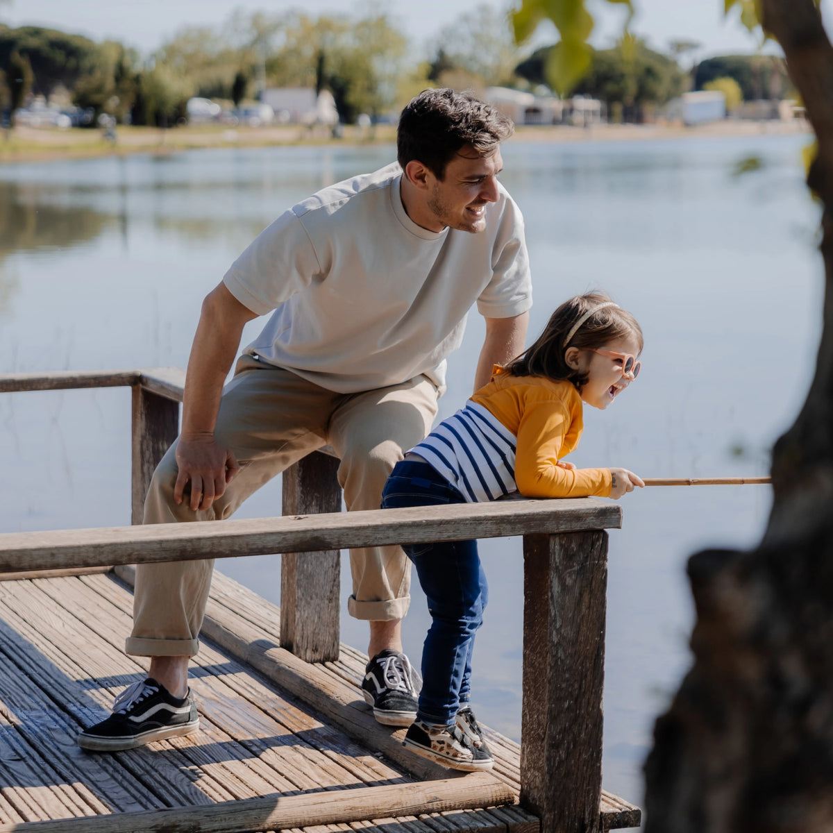 petite fille habillée avec le tee-shirt anti-noyade Floatee jaune à manches longues, sur un ponton avec une petite canne à pêche, et son papa à côté