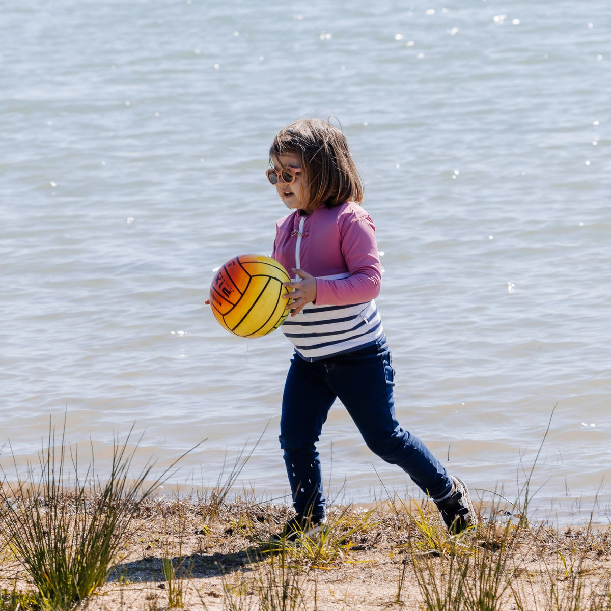 petite fille habillée avec le tee-shirt anti-noyade Floatee rose à manches longues, au bord d'un étang et portant un ballon
