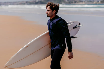 Homme sur une plage avec une planche de surf et portant le tee-shirt anti-noyade adulte manuel Floatee, manches longues, bleu