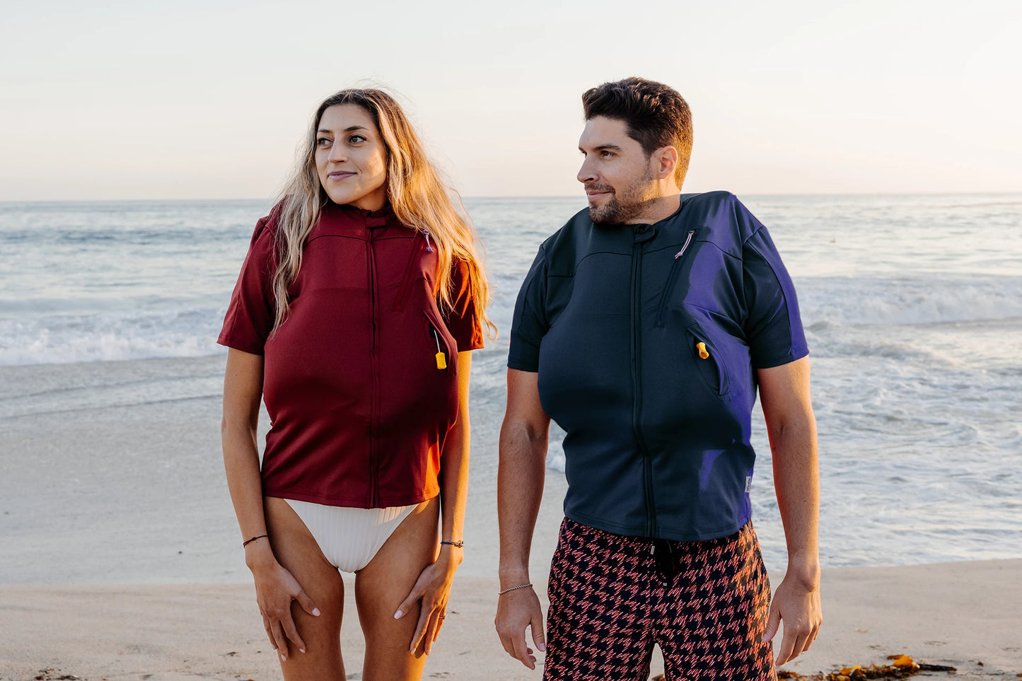 Homme et femme sur une plage et portant le tee-shirt anti-noyade adulte manuel Floatee gonflé, manches courtes, couleur bleue pour l'homme et rouge pour la femme