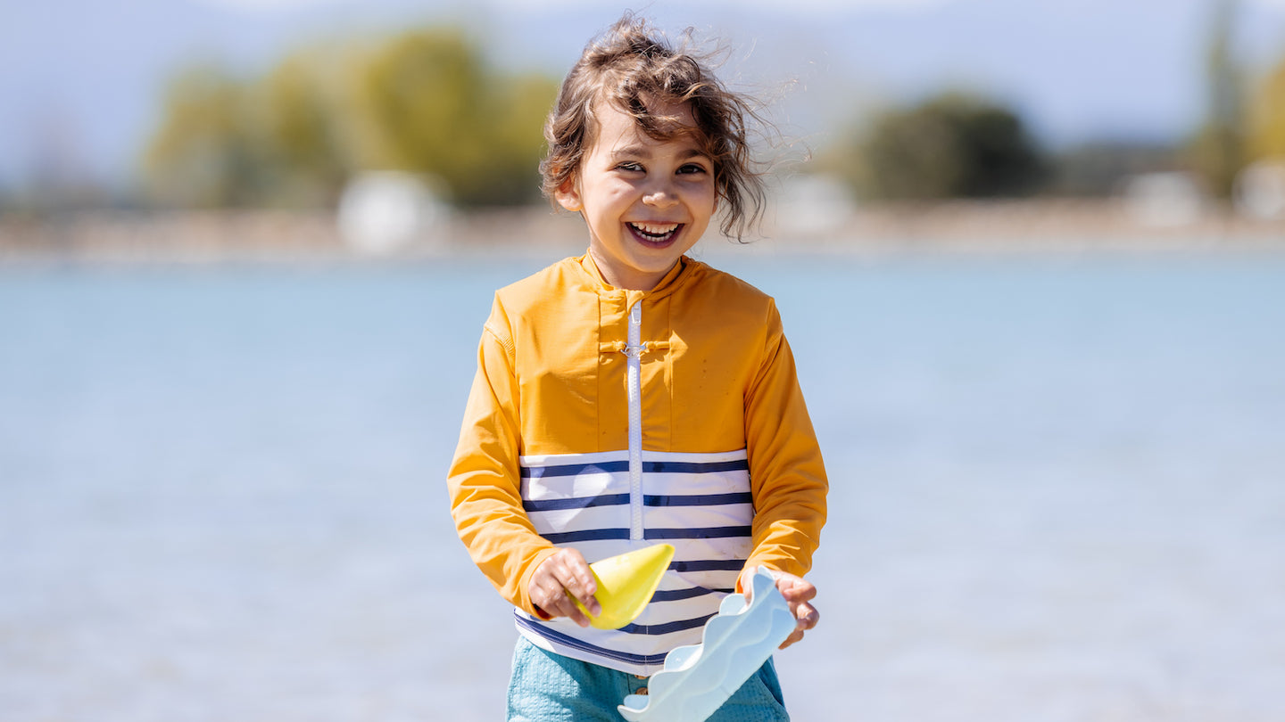 petite fille vue de face portant le Tee-shirt anti-noyade pour enfant Floatee, haut jaune et bas marinière, tenant dans ses mains des jouets